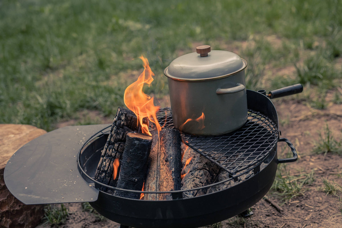 Outdoor enamel stock pot on a fire pit grill with flames, lid closed, dark gray