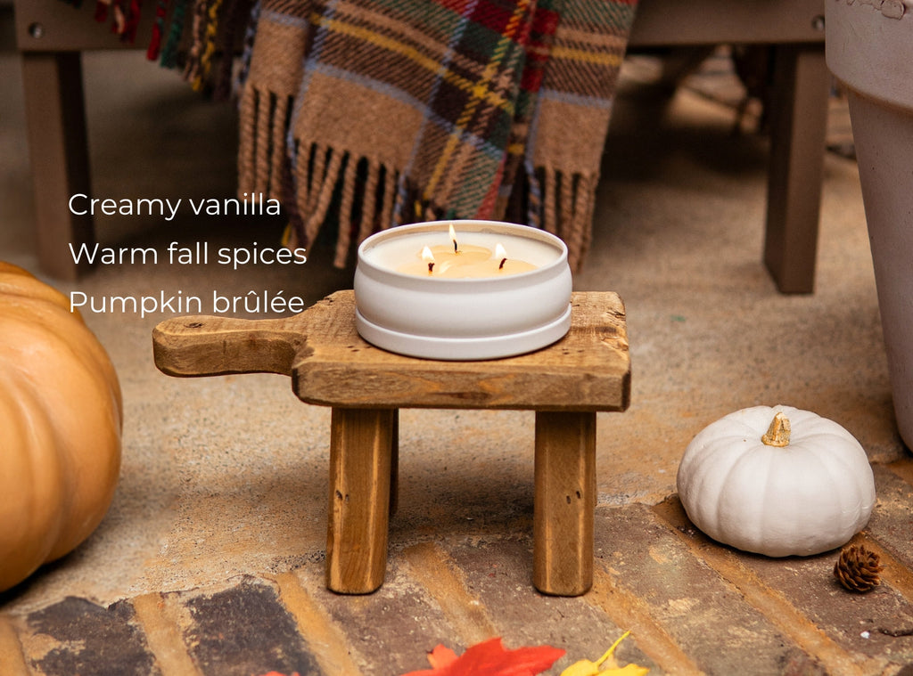 Lit white 3-wick candle in a round tin on a small wooden stool with pumpkins