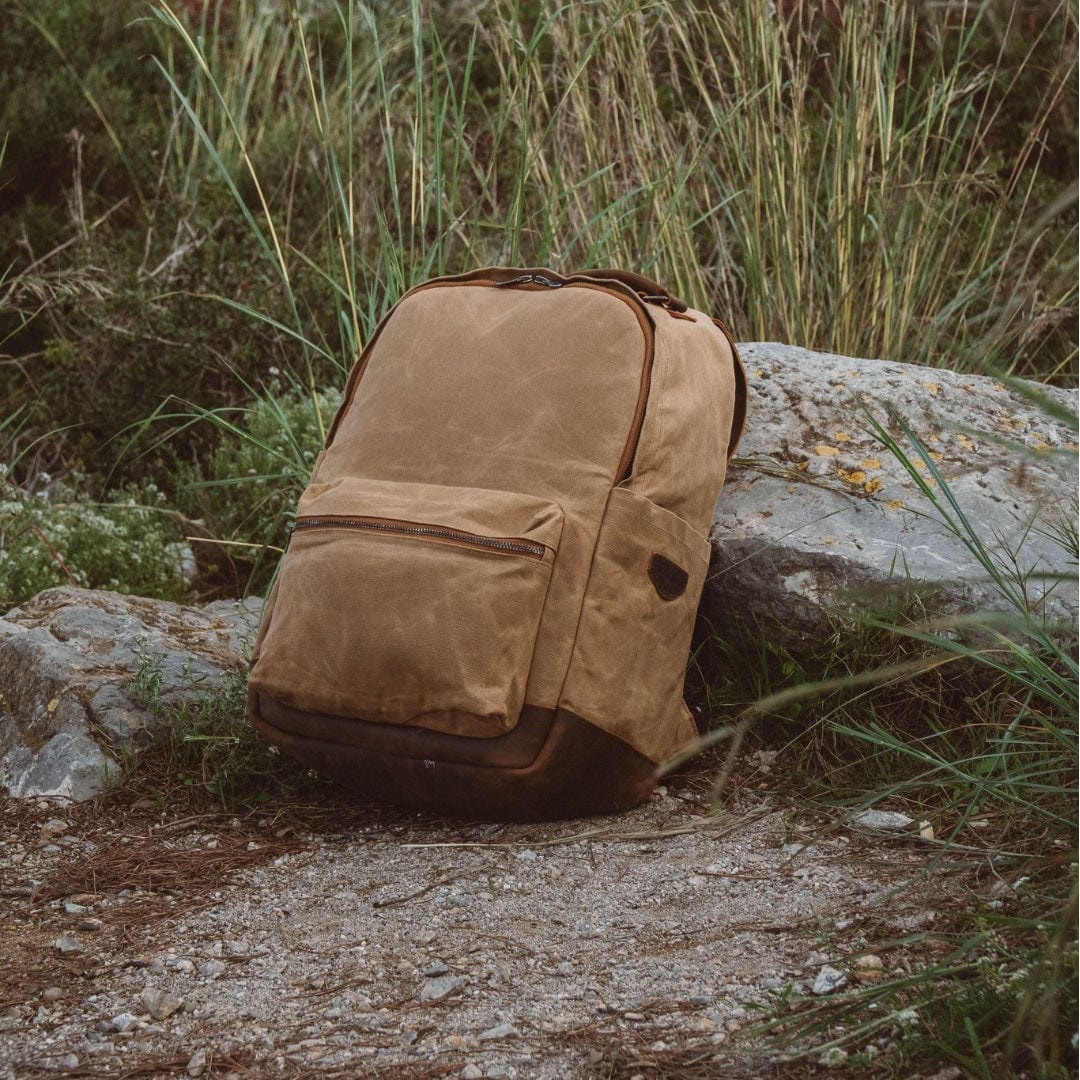 Sand waxed canvas backpack resting on rocks with front pocket and leather trim