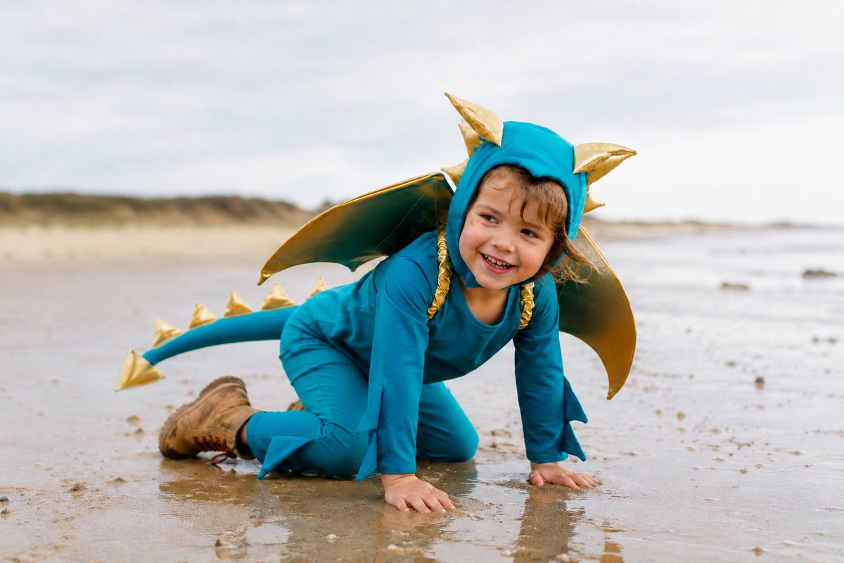 Turquoise dragon costume with gold wing headpiece and cape, toddler crouching outdoors.