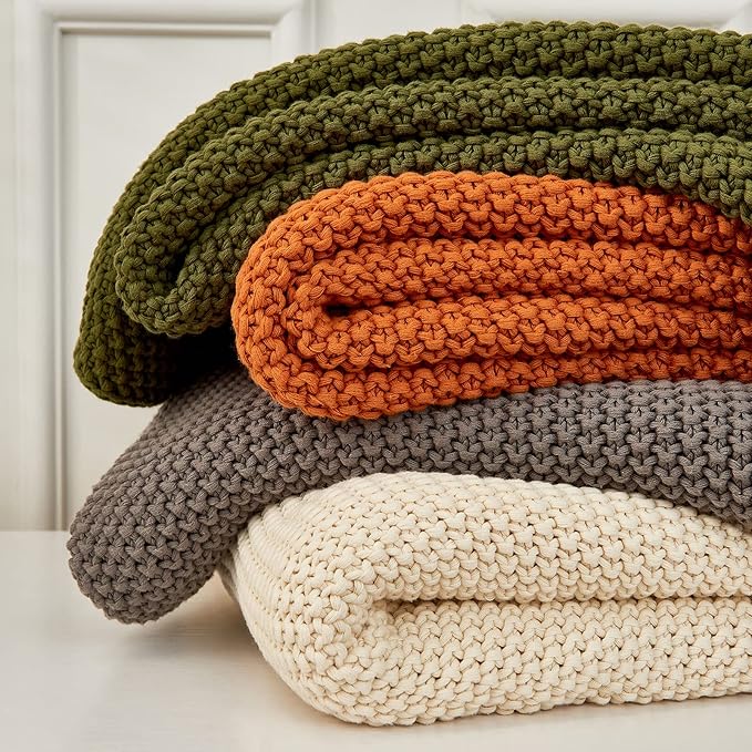 Stacked chunky knit throw blankets in olive green, orange, and cream on a sofa.