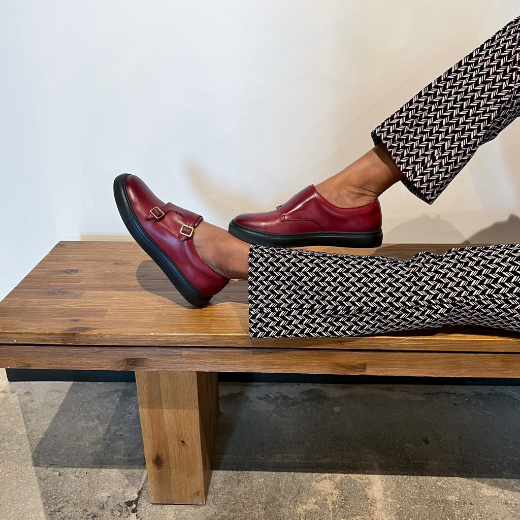 Women’s dark red slip-on shoes with gold buckle straps, styled on a wooden bench