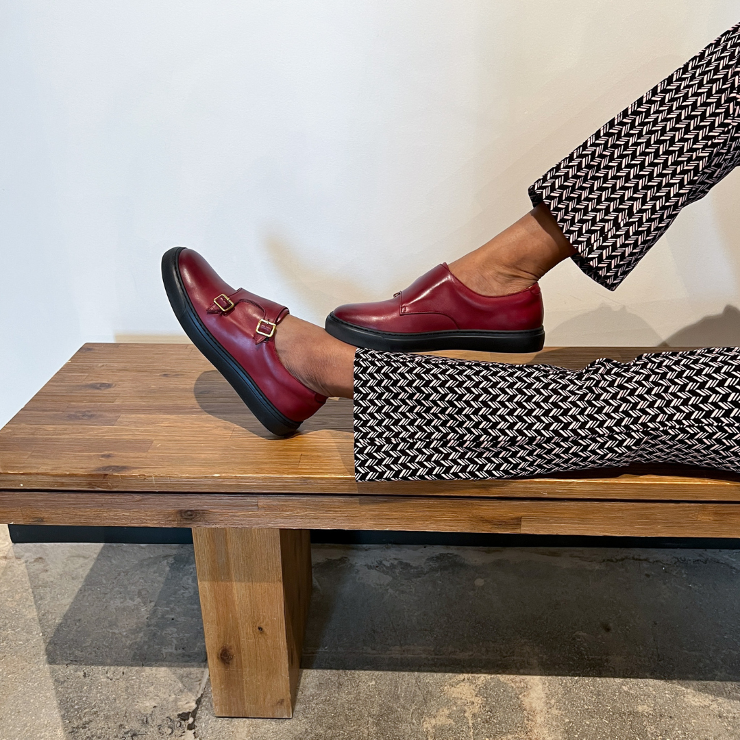 Women’s dark red slip-on shoes with gold buckle straps, styled on a wooden bench