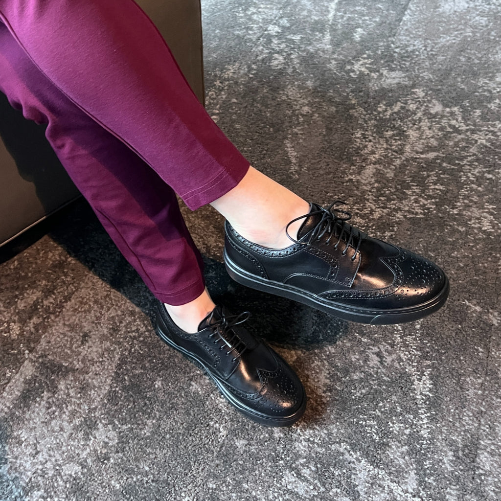 Black lace-up oxford shoes on feet with perforated detailing and flat sole