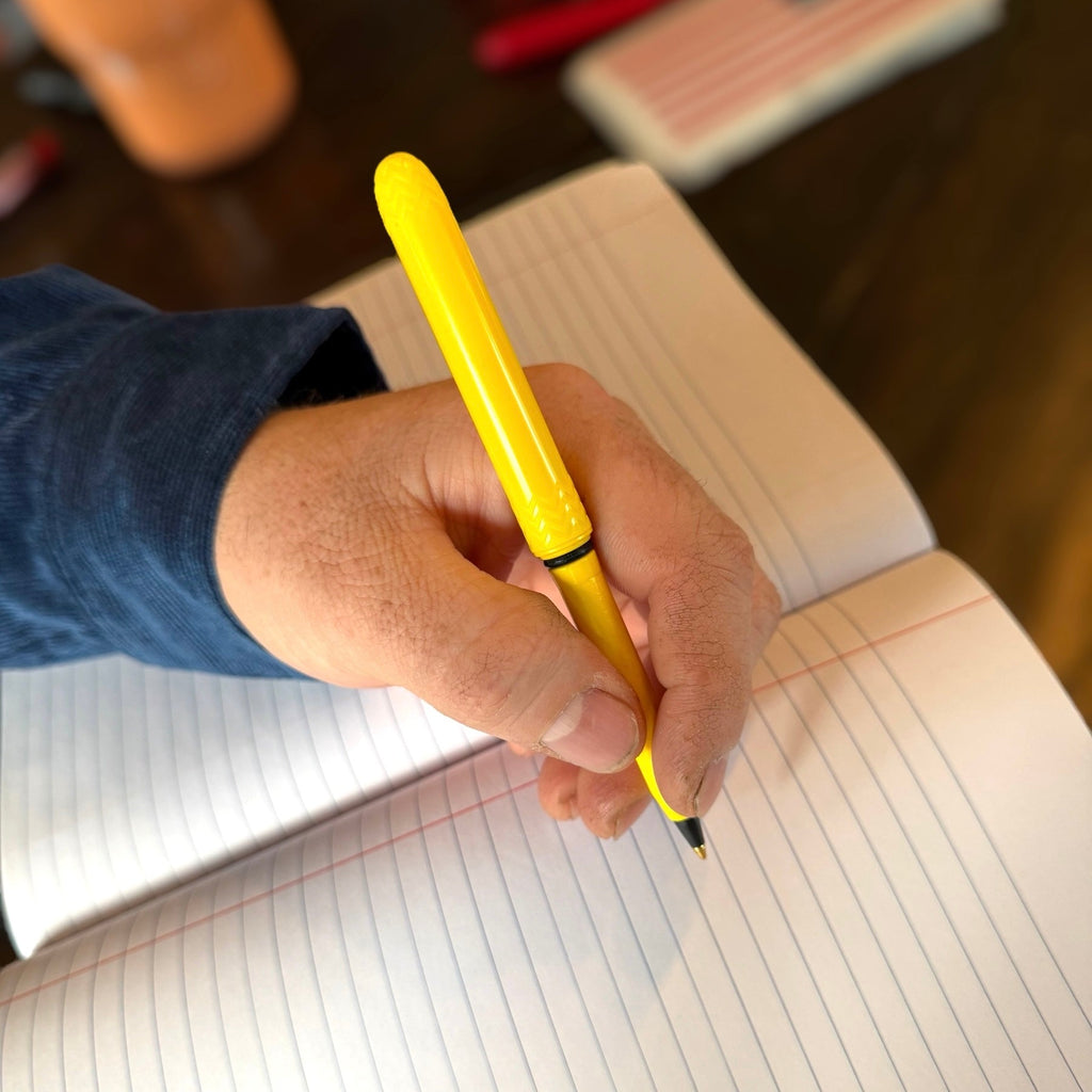 Yellow pocket pen with zigzag grip, held by hand writing in lined notebook.
