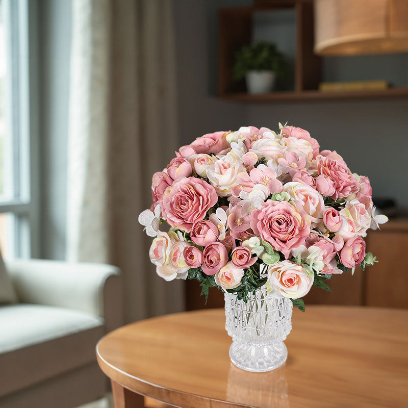 Pink silk rose bouquet with small white blossoms and green leaves in a clear vase