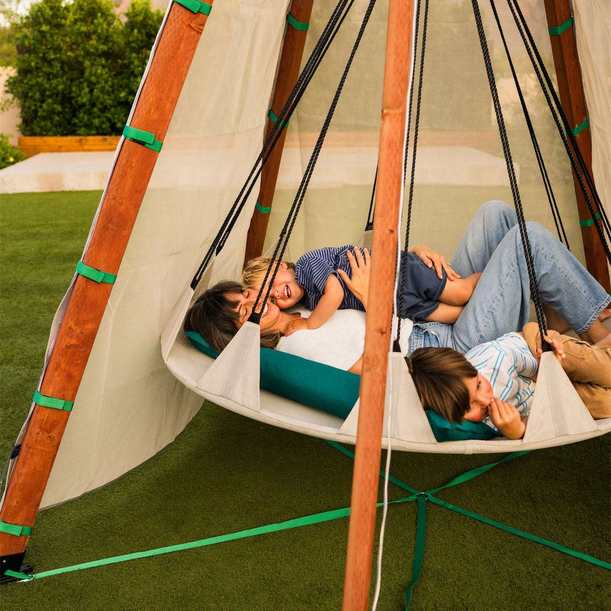 Off-white hanging tent bed with two kids relaxing inside; wooden poles and green trim