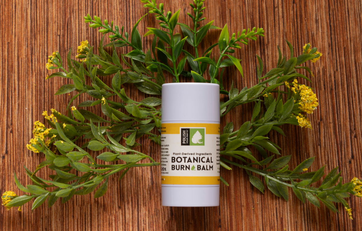 Botanical balm stick in a white container with yellow label and green foliage