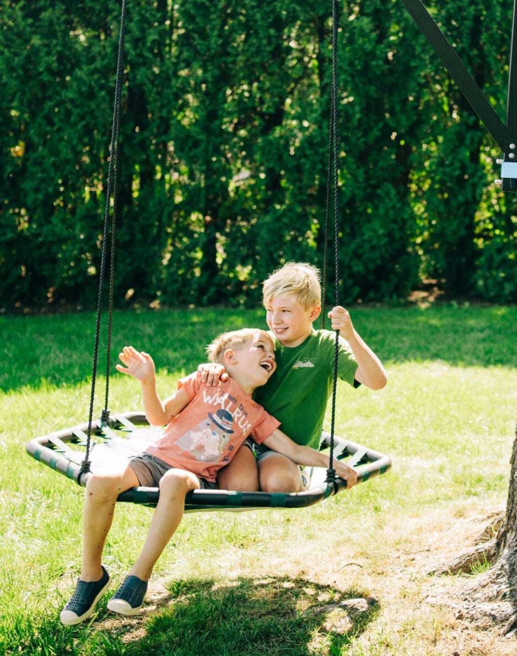 Two children playing on a green rope swing with woven seat and hanging cords