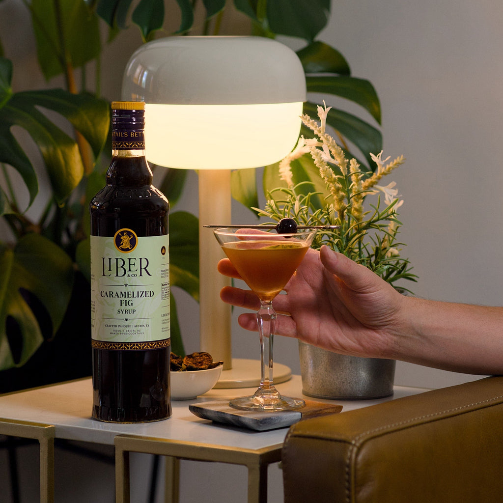 Caramelized fig syrup bottle beside a cocktail glass held by a hand on a tabletop