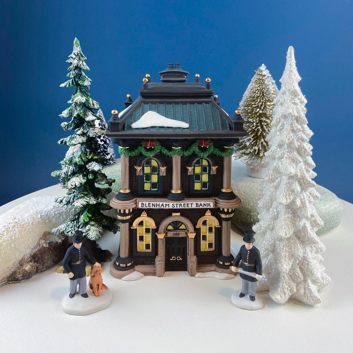 Illuminated bank house with snow roof, two policemen figurines, and small dog