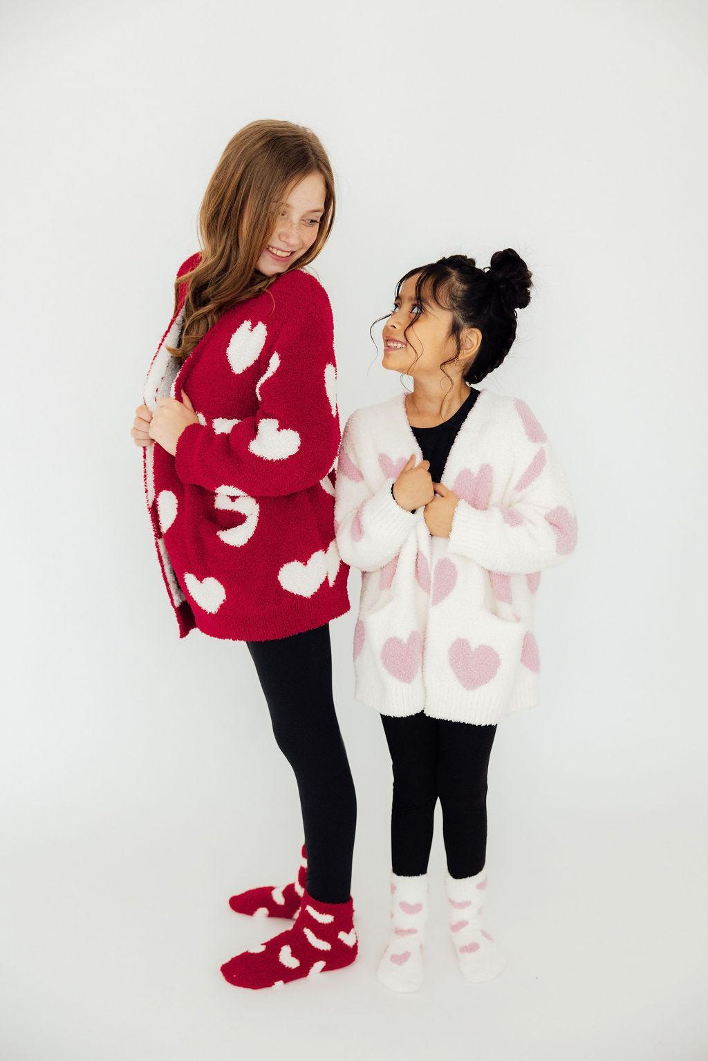 Light pink heart cozy cardigan on child, fuzzy white knit with large heart print and long sleeves.