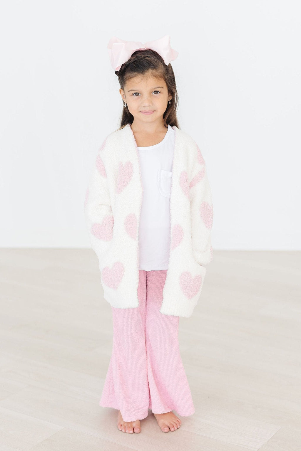 Light pink heart cozy cardigan worn by child, fuzzy white knit with heart print and long sleeves.