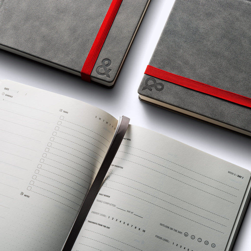 Open planner pages beside two gray covers with red ribbon bands and ampersand