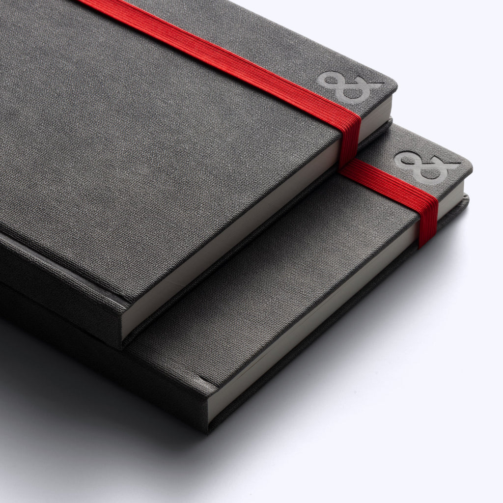 Two stacked gray habit system planners with red ribbon bands and embossed ampersand