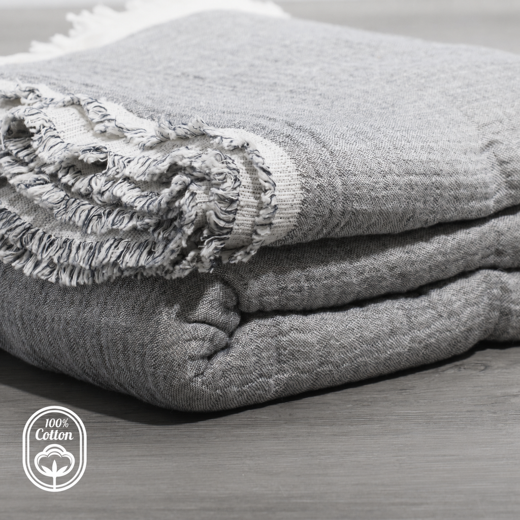 Folded grey reversible muslin coverlet with fringed trim and layered quilting texture