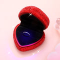 Heart-shaped ring case with red rhinestone exterior, open lid revealing blue velvet insert