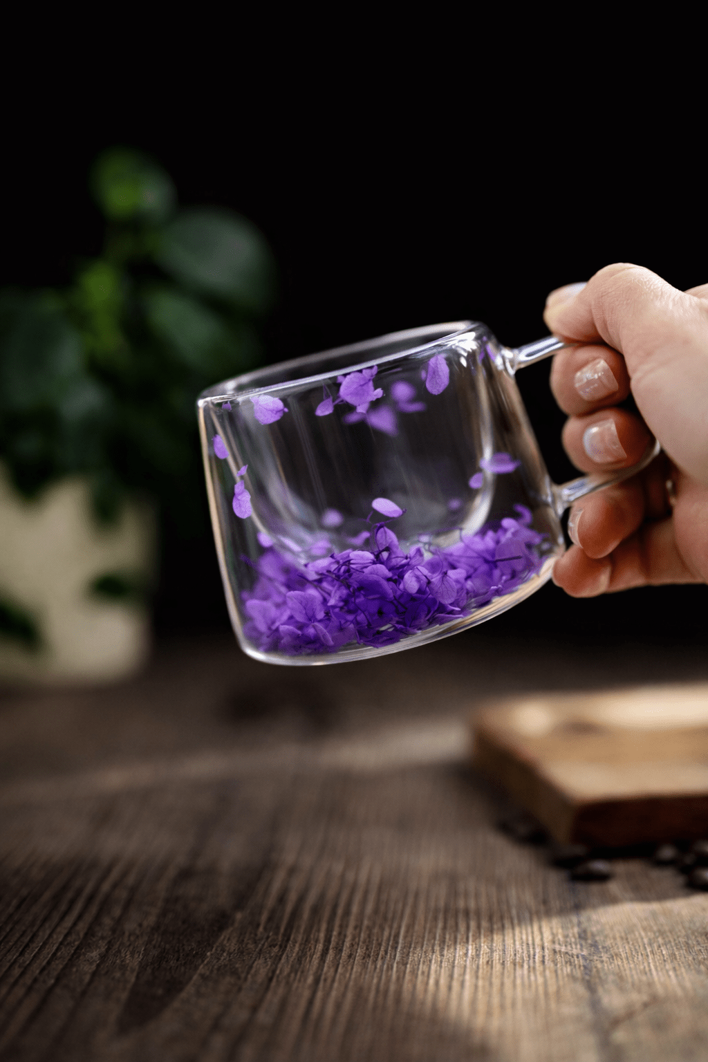 Hand holding floral glass mug with purple dried flowers, clear handle, filled with dark tea