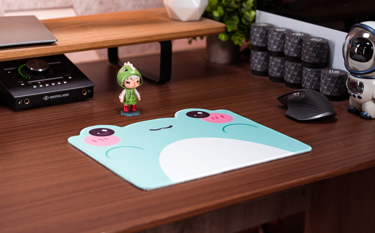 Teal cartoon frog desk mat with white belly and pink cheeks, placed on desk surface