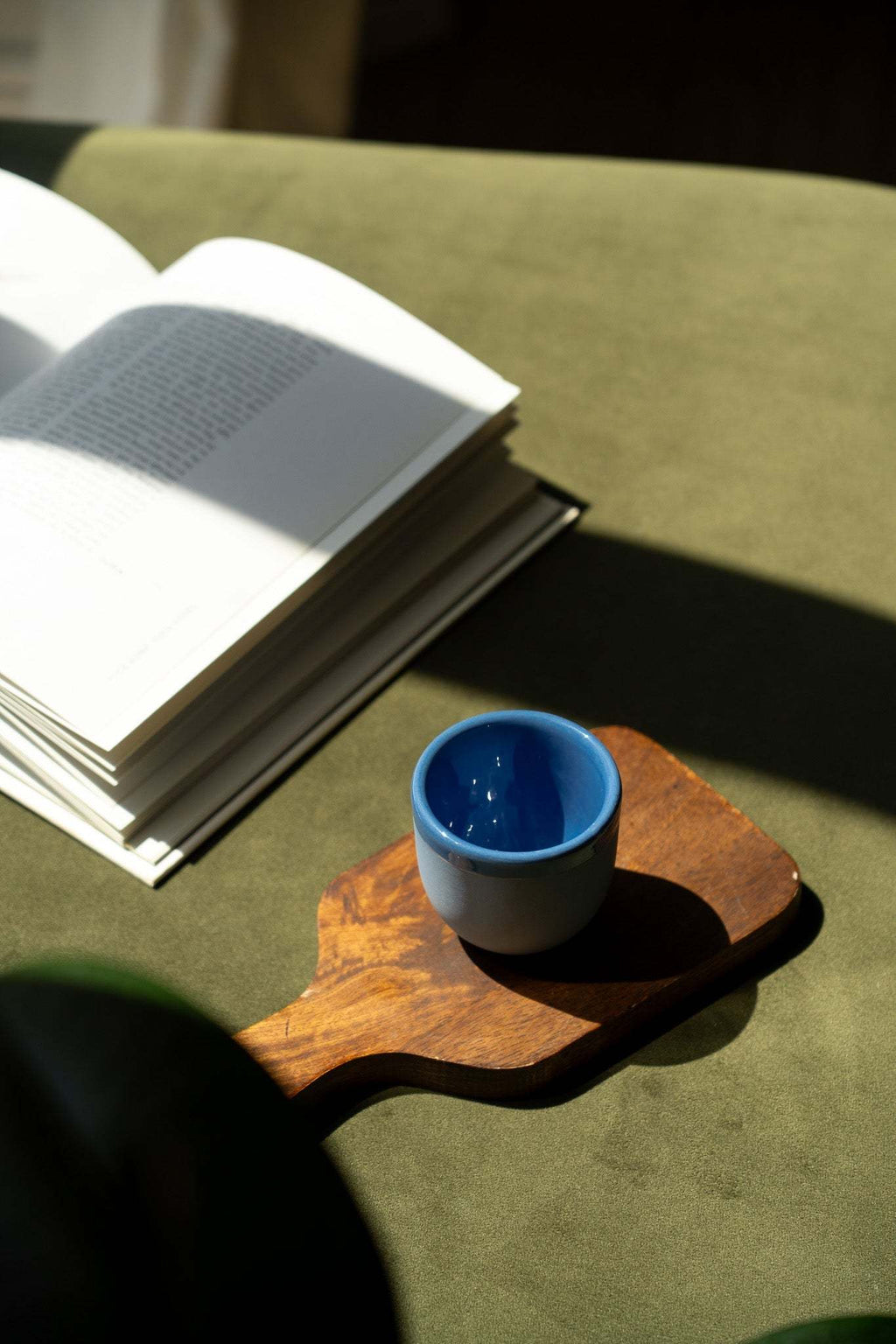 Handmade blue porcelain espresso cup on wooden tray, beside an open book
