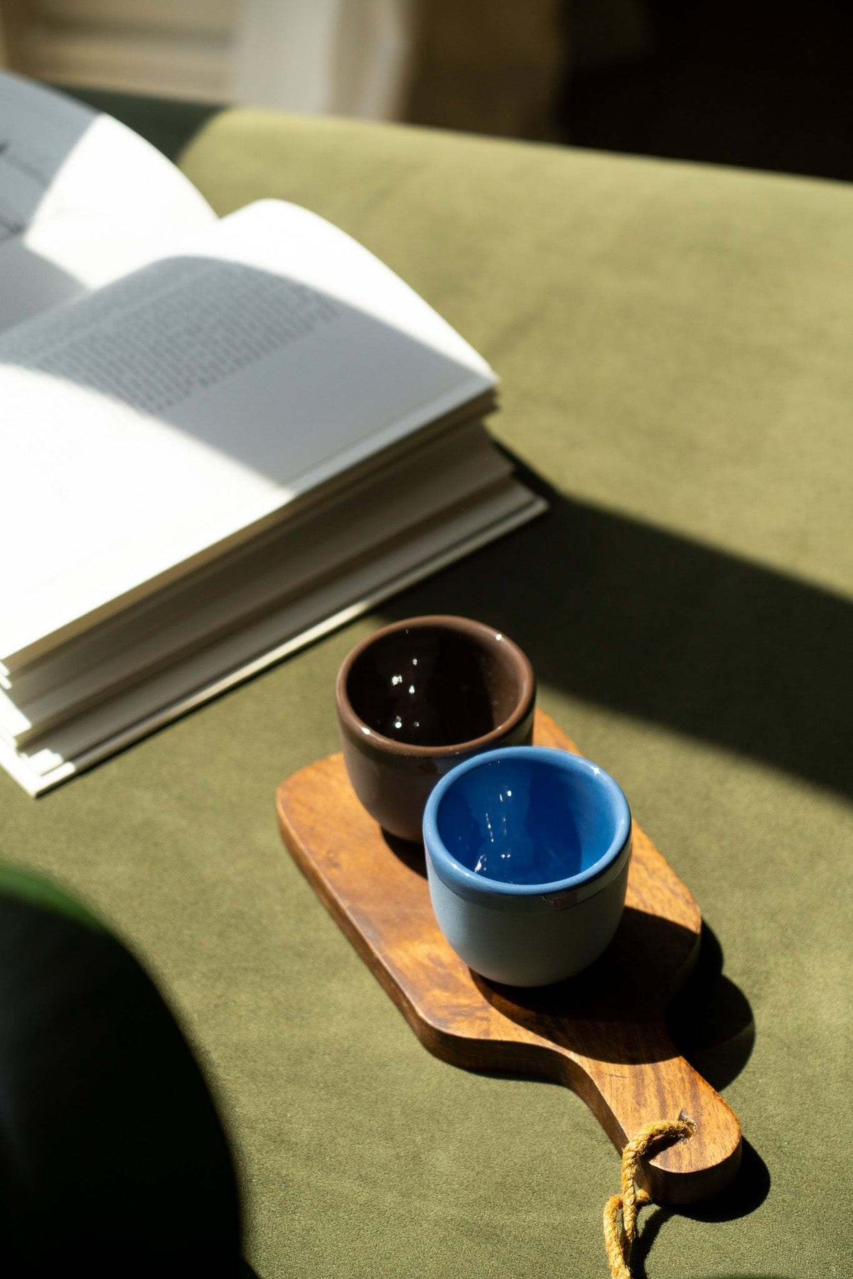 Brown and blue ceramic espresso cups on a wooden tray beside an open book.