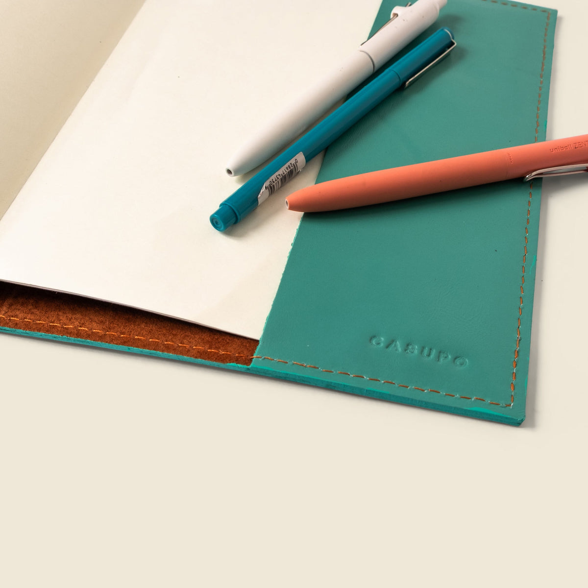 Teal leather notebook cover with embossed logo and stitched trim, with pens on paper.