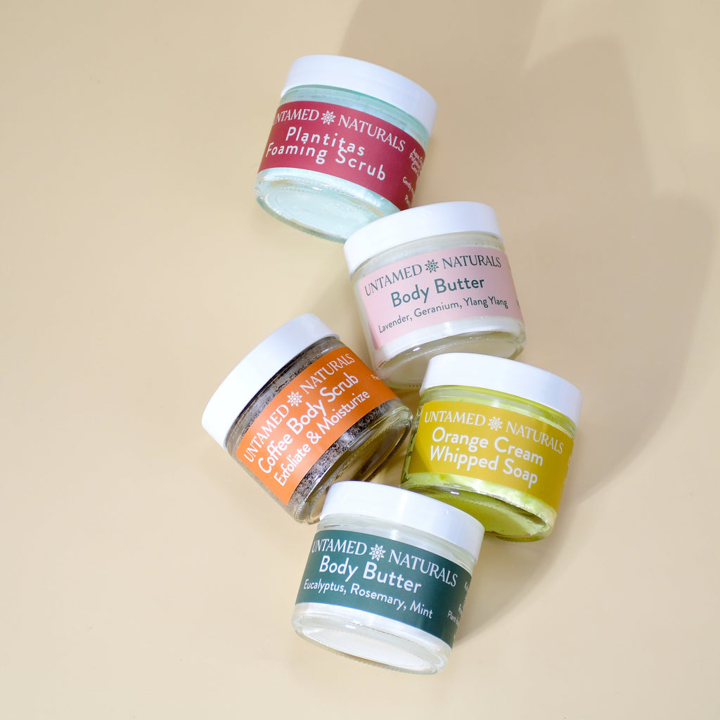 Stacked body care jars with labels: body butter, coffee scrub, orange whipped soap, foaming scrub