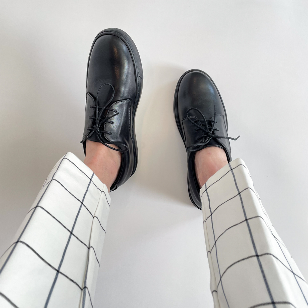 Black lace-up oxfords on feet with rounded toe and thin laces; worn with plaid pants.