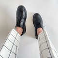 Black lace-up oxfords on feet with rounded toe and thin laces; worn with plaid pants.