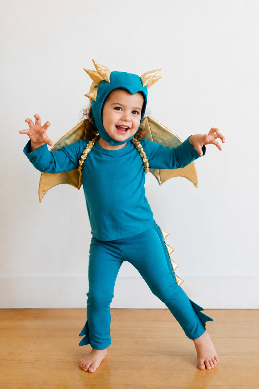 Turquoise dragon costume with gold wing headpiece and cape, toddler arms out indoors.
