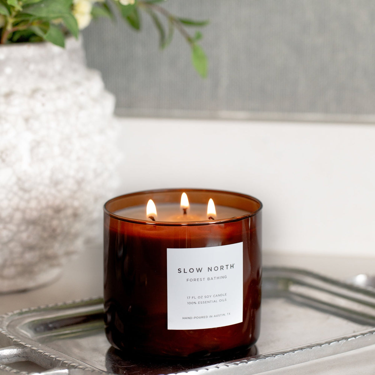 Amber glass 3-wick candle with three lit flames and white label on metal tray