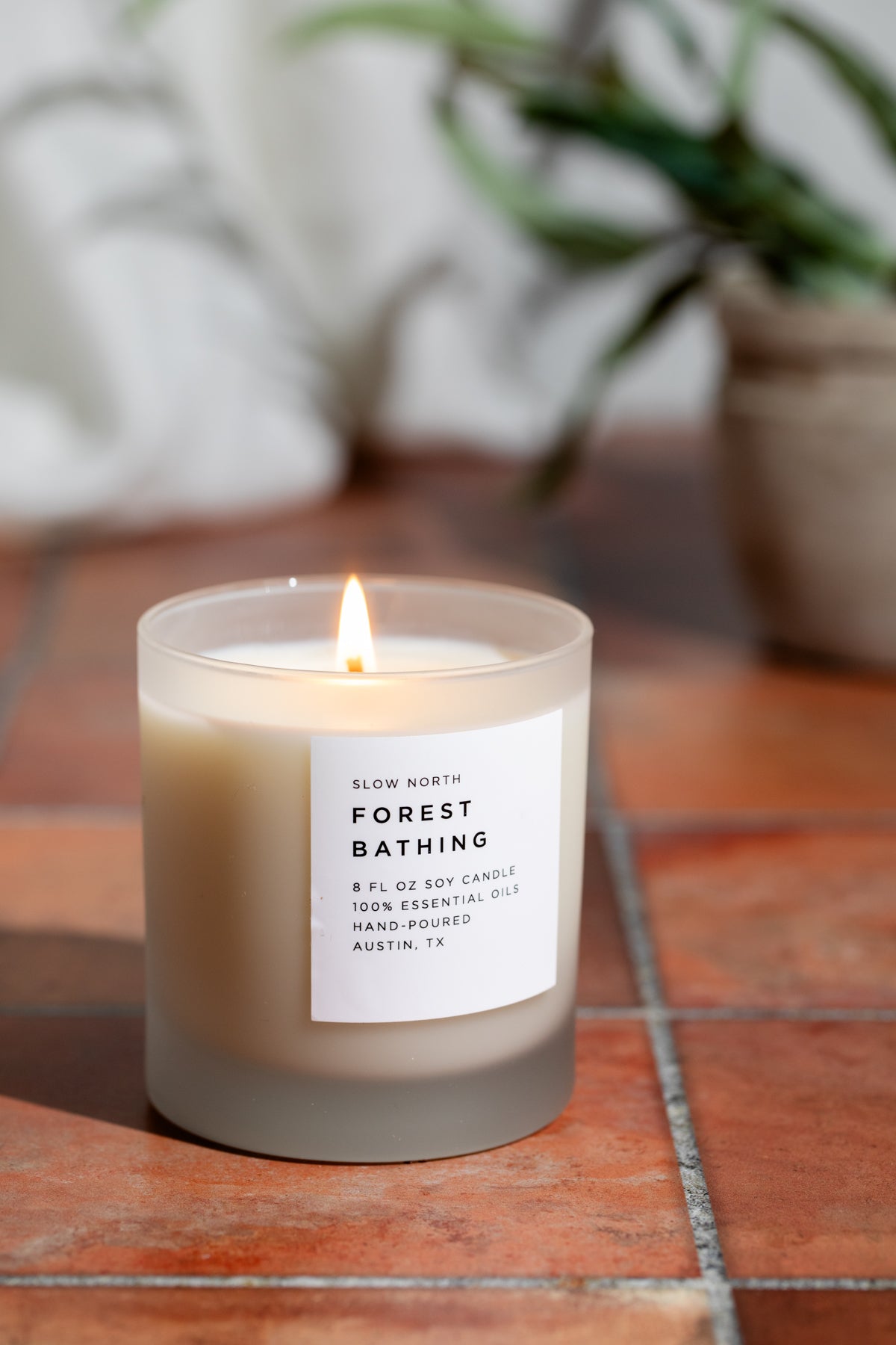 Soy candle in frosted jar with lit wick and white label on terracotta tile