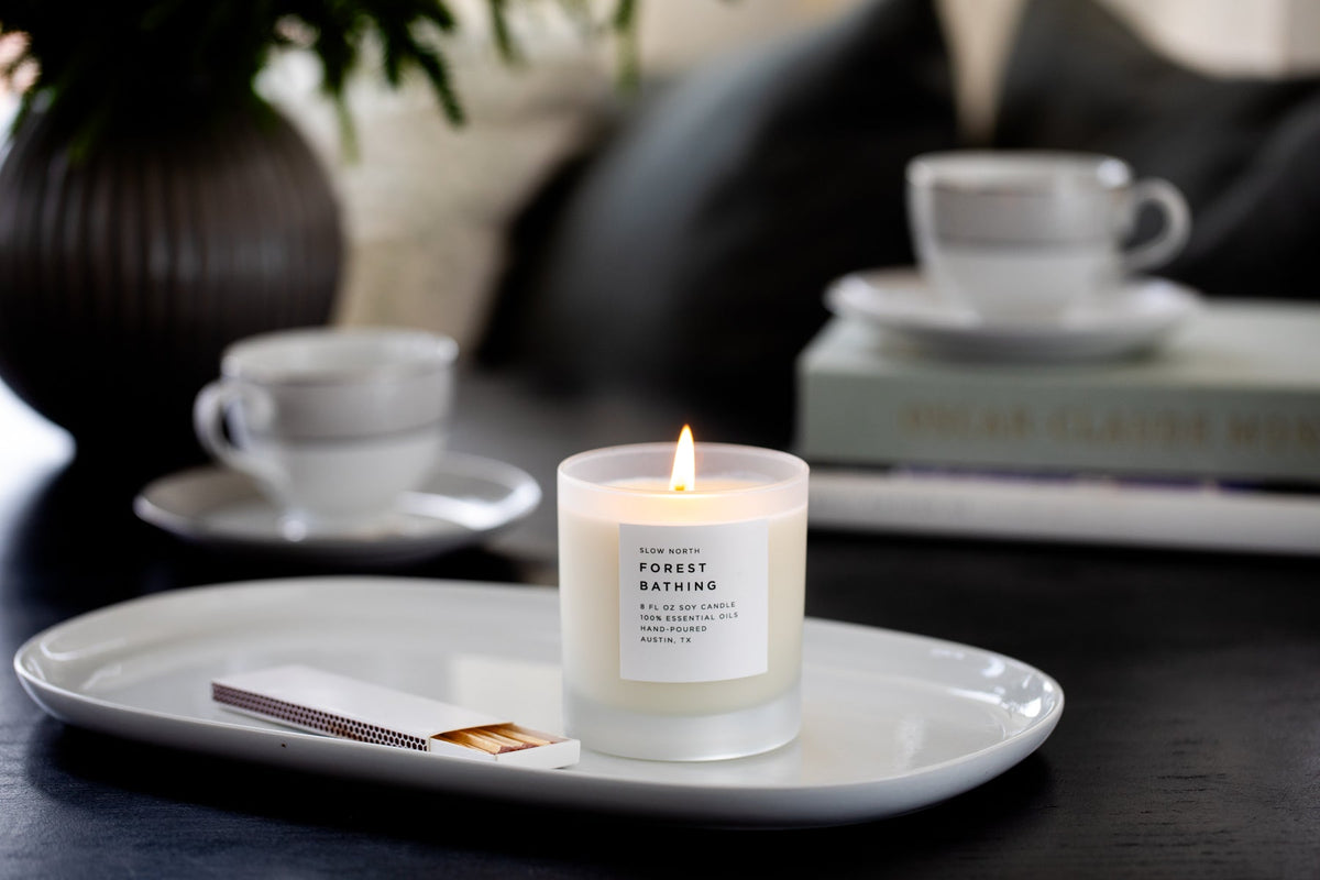 Soy candle in frosted jar with lit wick on tray, with nearby coffee cups
