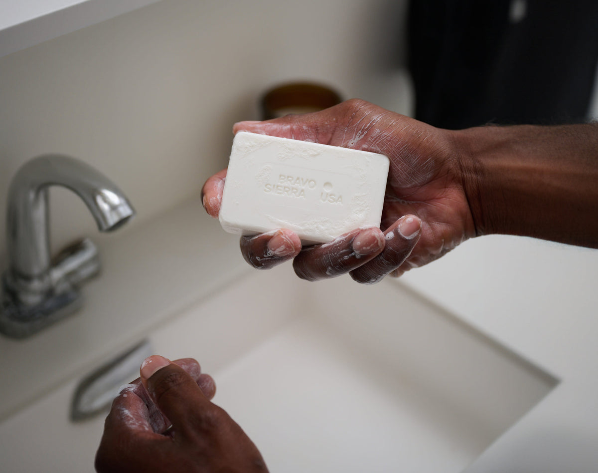 Hands holding a beige face and body cleansing bar soap over sink