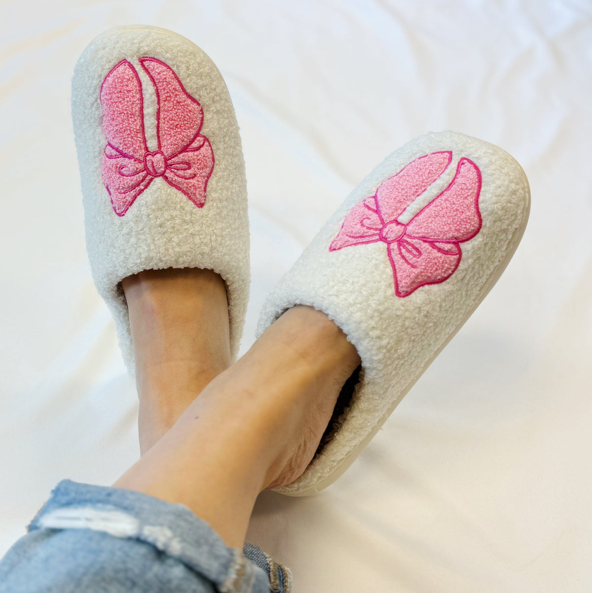 Cream lounge slippers with pink bow embroidery on top, worn with jeans