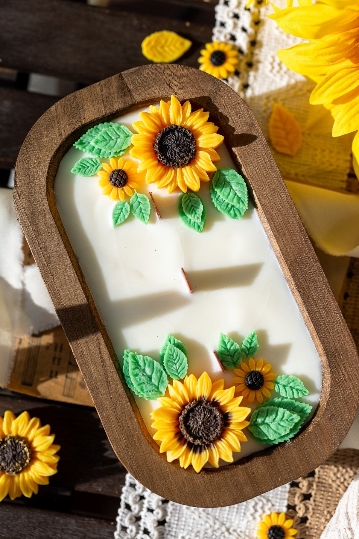 Angled view wooden sunflower soy wax candle with yellow blooms, green leaves, white ribbon bow, red wick