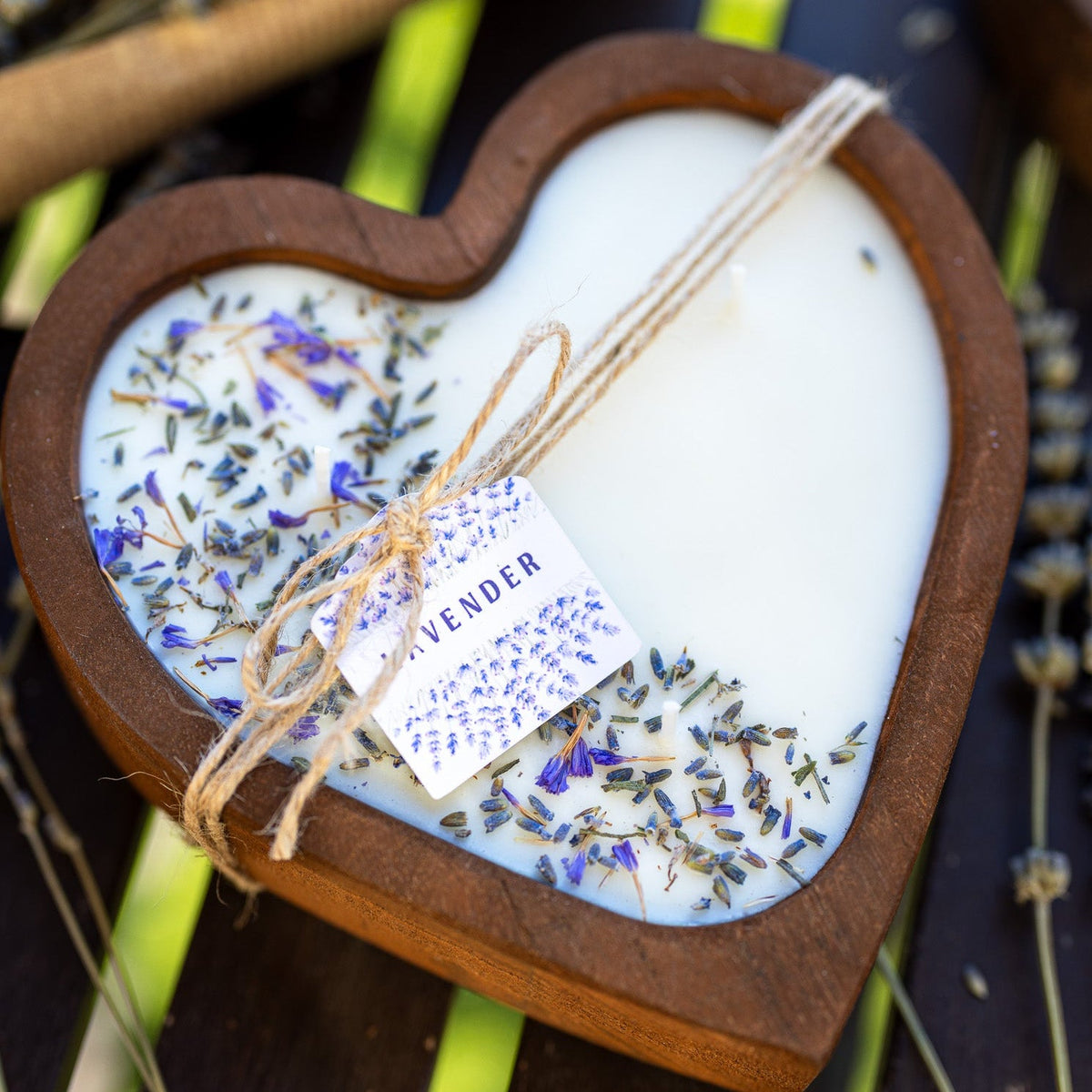 Heart-shaped wooden bowl candle with white wax, blue-purple lavender bits, and hanging label.