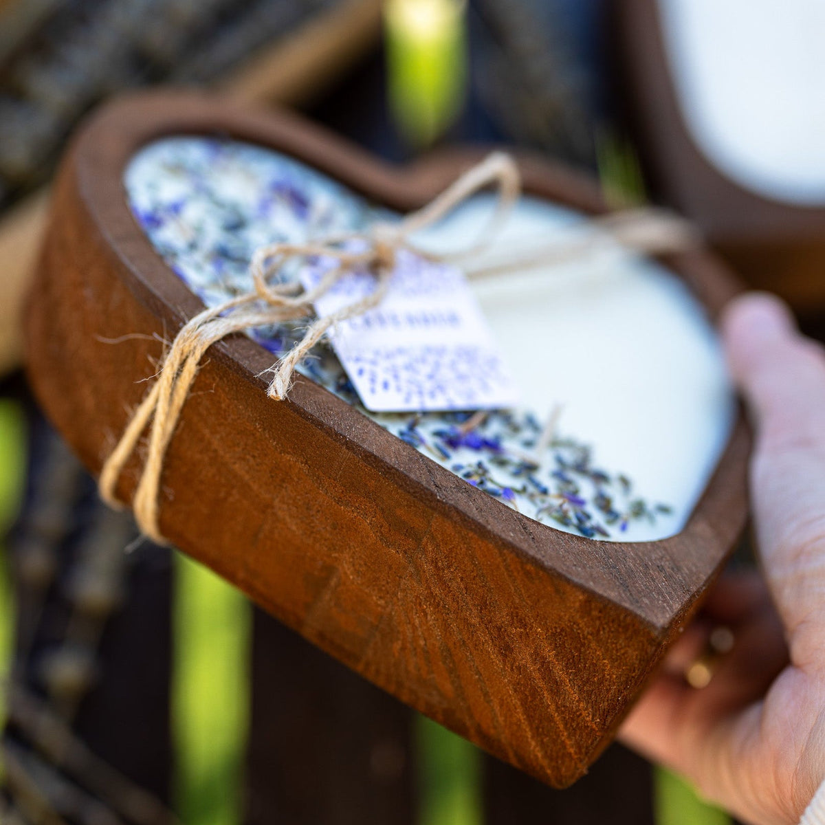 Hand holding heart-shaped wooden bowl candle with white wax and lavender sprigs visible.