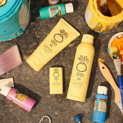 Kids SPF 50 sunscreen products in yellow tubes and bottle on a beach bag setup