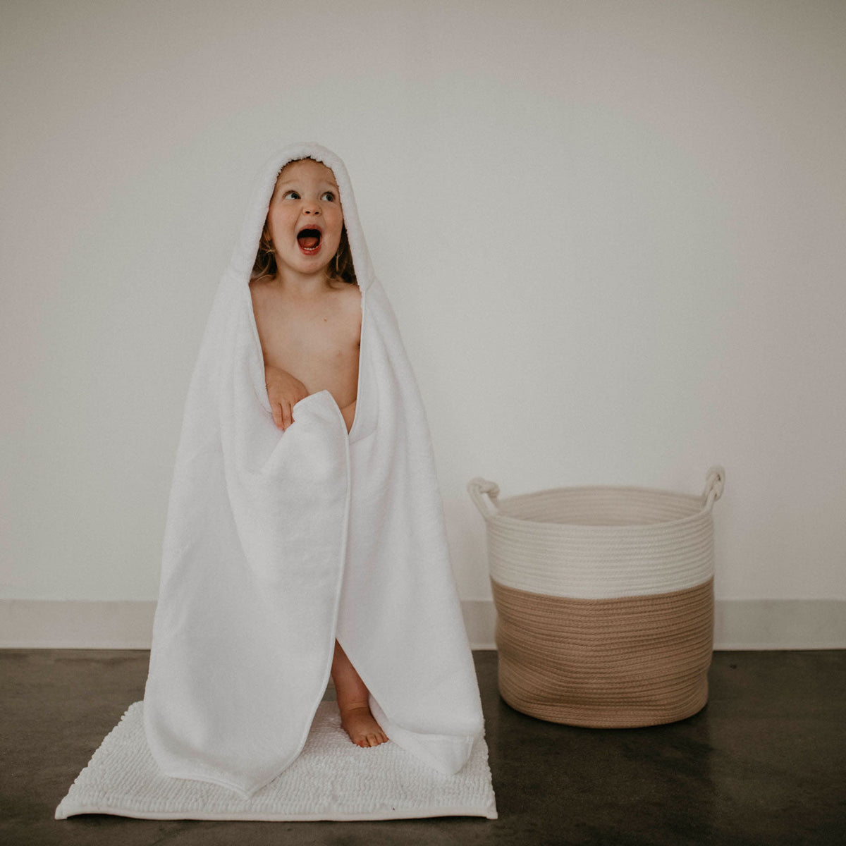 White hooded bath towel with baby standing, robe draped over shoulders
