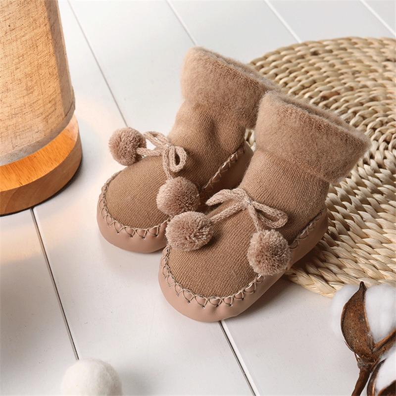 Newborn baby shoes in tan fabric with bow and pom pom details on the ankle