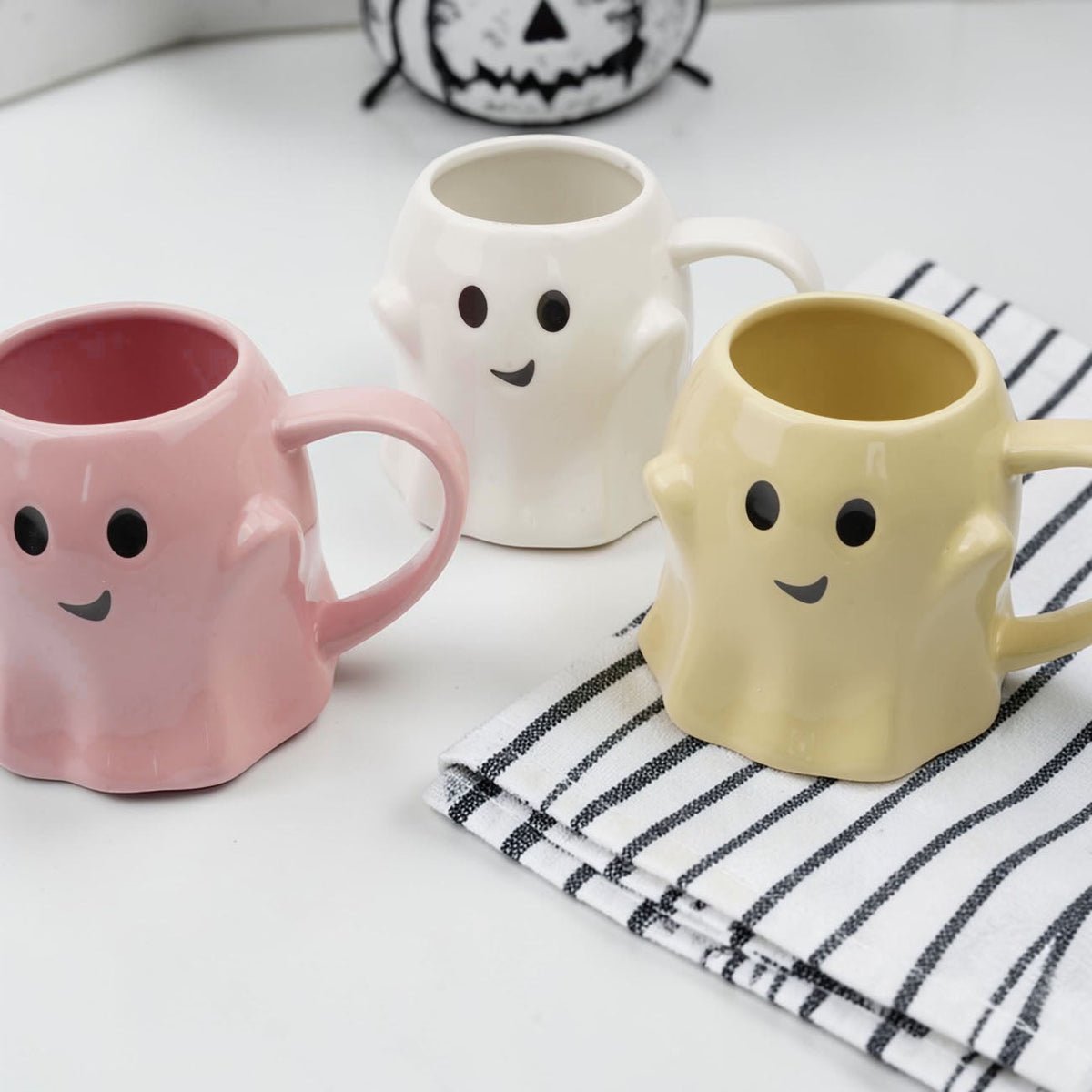 Three ghost-shaped ceramic mugs in pink, white, and yellow with smile faces