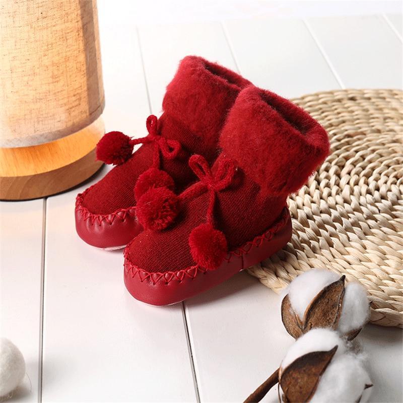 Newborn baby shoes in red plush with bow and pom pom details on the ankle