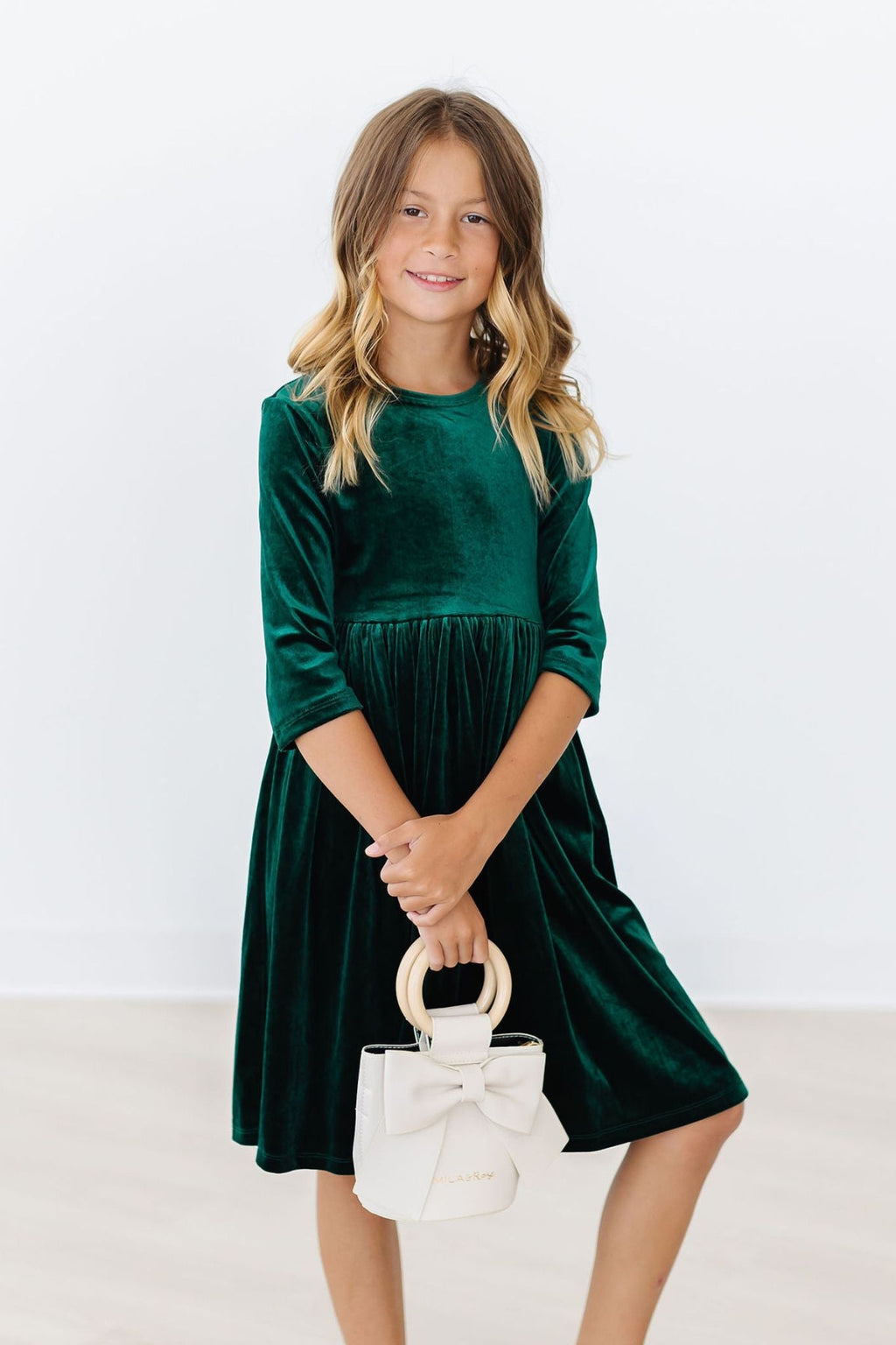 Dark green velvet twirl dress with 3/4 sleeves and gathered skirt, child holding a bag