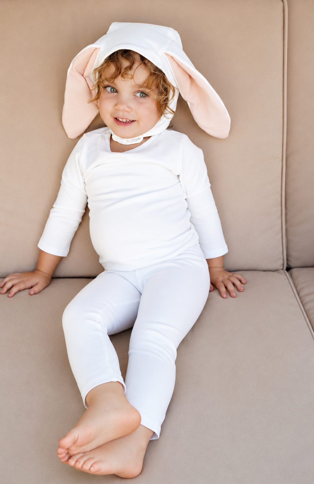 Baby ivory bunny hooded romper with long pink ears, sitting on sofa