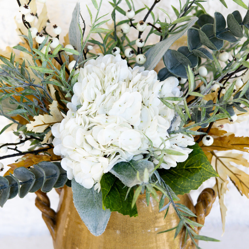 Floral bouquet in gold vase close-up with dense white flowers, eucalyptus leaves, and pods