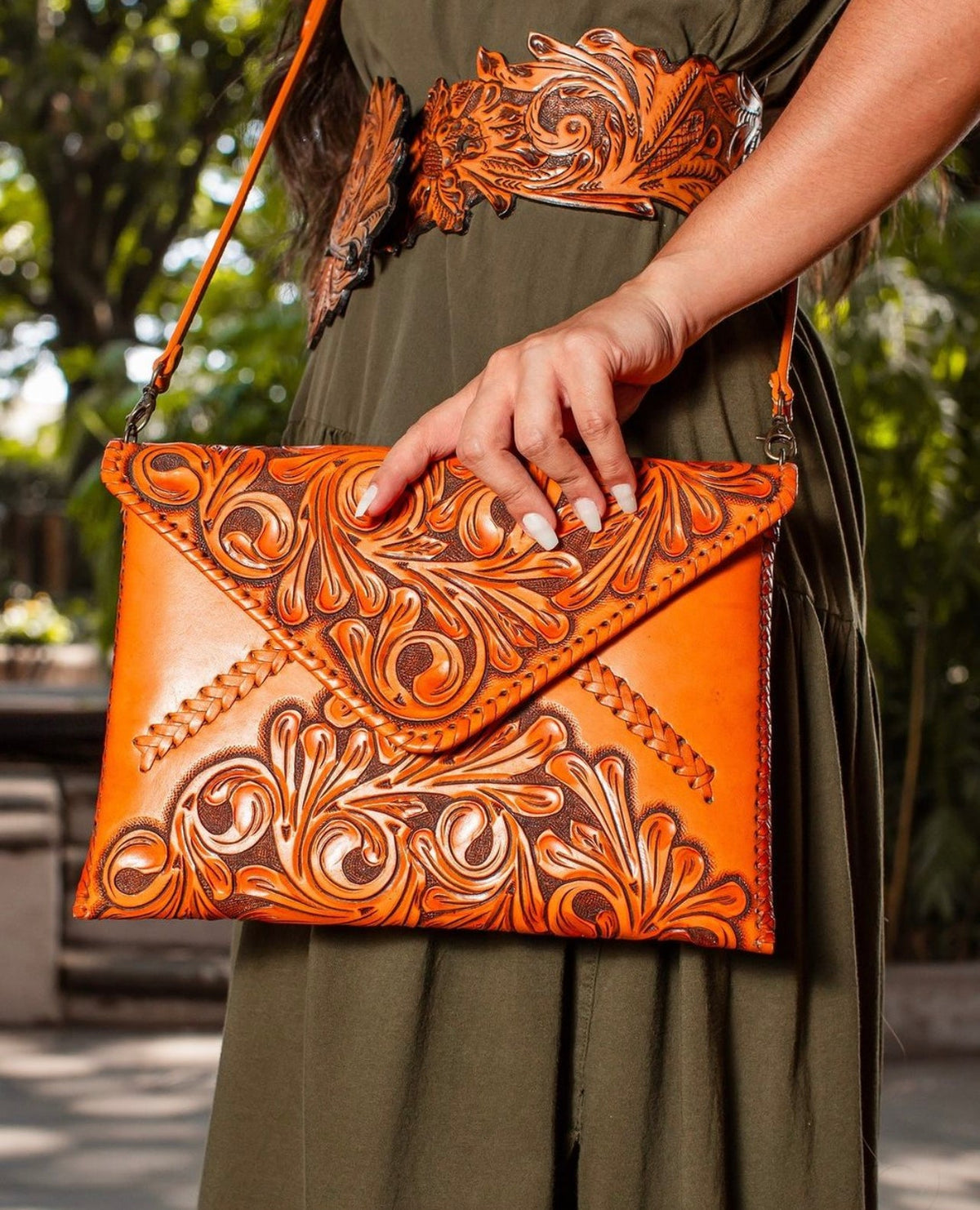 Hand-tooled leather crossbody clutch bag held by model, orange with carved floral