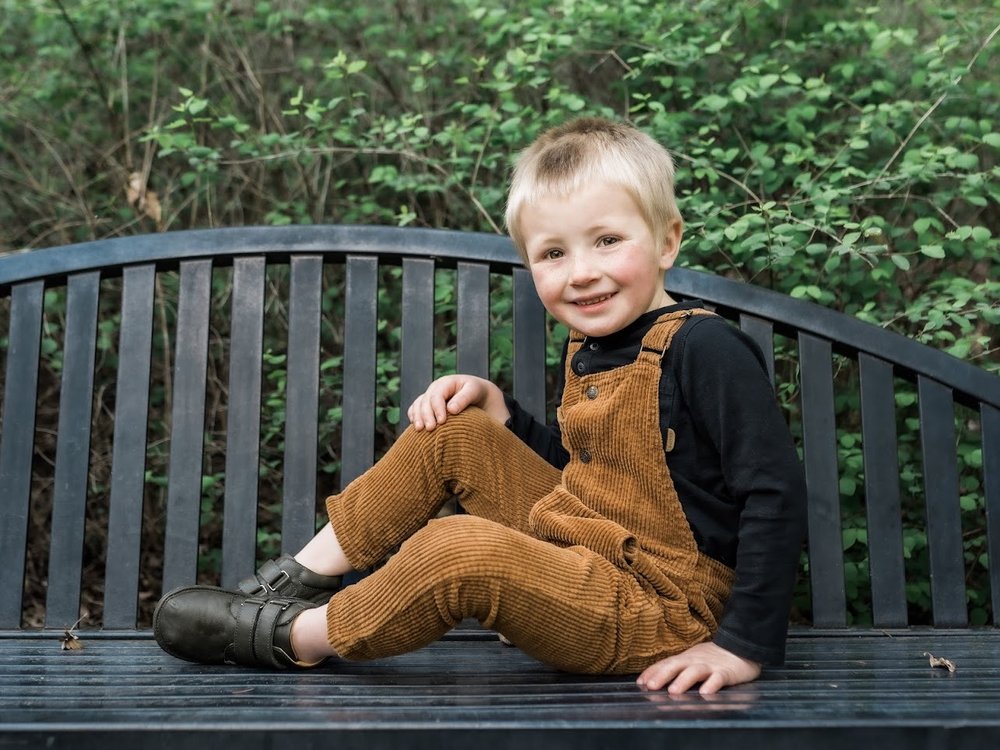 Toddler barefoot sneakers, dark green, worn with brown overalls on a bench outdoors.