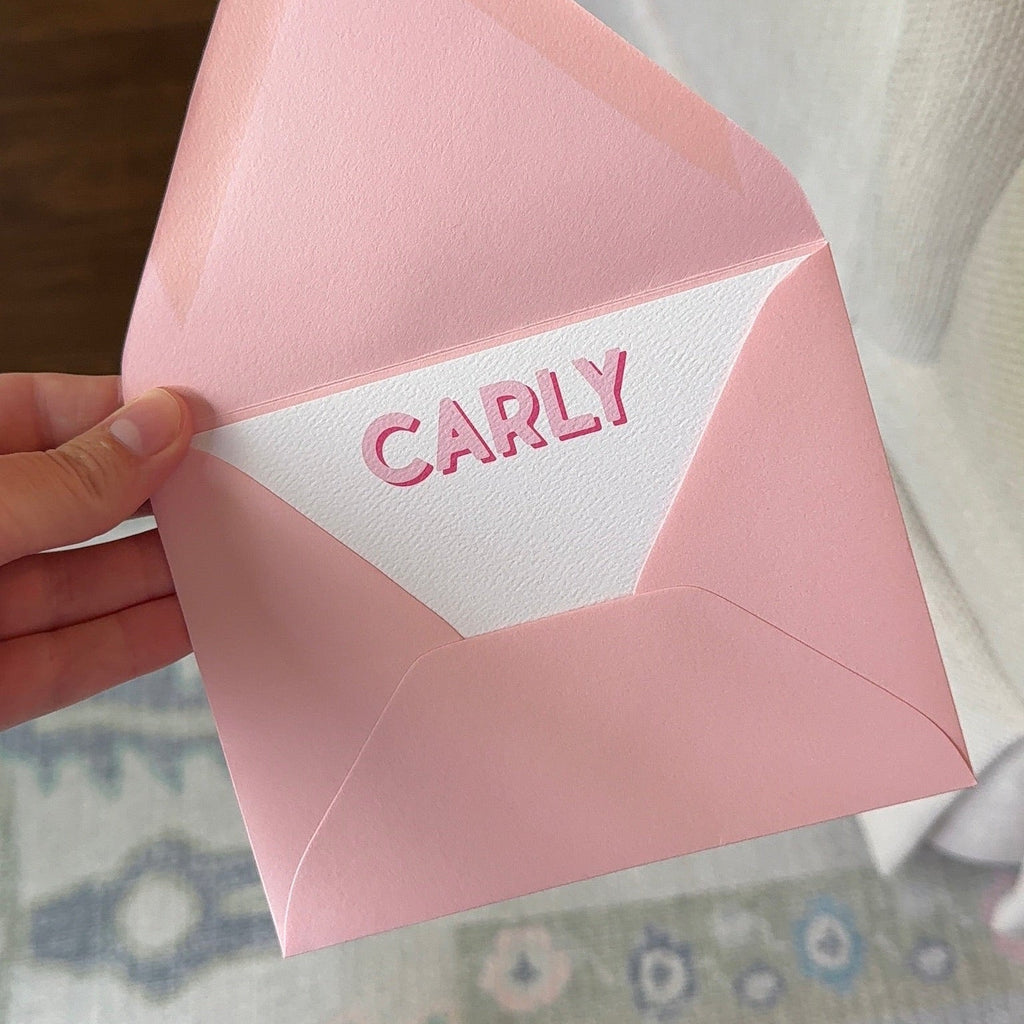Hand holding a pink envelope with “CARLY” printed on a white address card