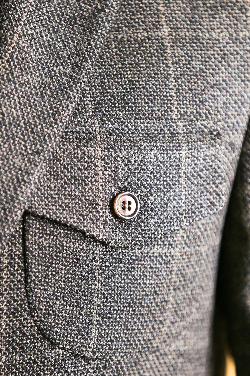 Men's blazer pocket flap detail in dark gray textured fabric with metal button.
